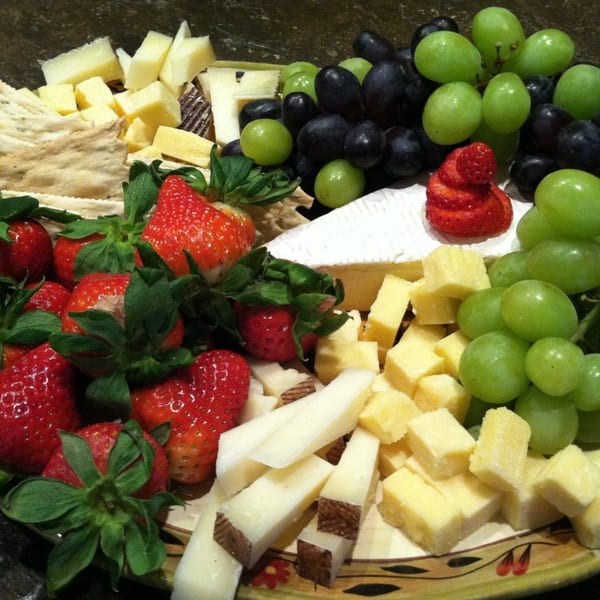 A platter of cheeses, crackers, strawberries, and green and black grapes.