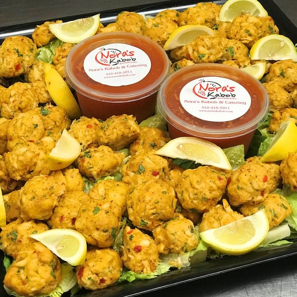 A catering tray of Noras Kabob's Mediterranean crab cakes with lemon wedges and two sauce containers.