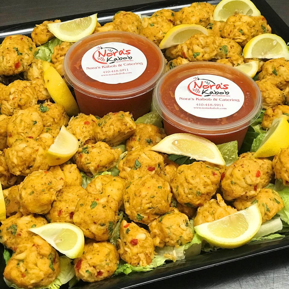 A catering tray of Noras Kabob's Mediterranean crab cakes with lemon wedges and two sauce containers.
