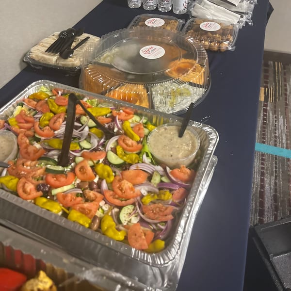 A catering spread from Noras Kabob featuring a large Mediterranean salad, pita bread, and desserts.