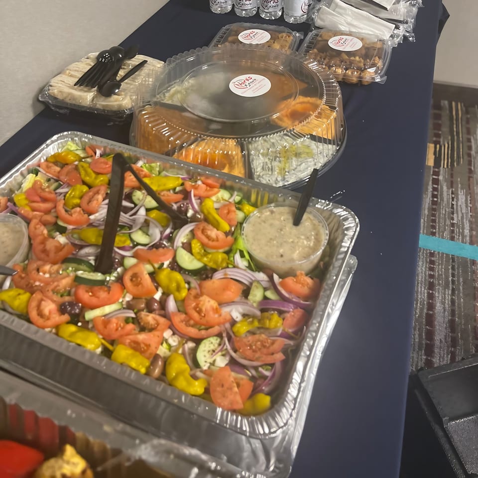 A catering spread from Noras Kabob featuring a large Mediterranean salad, pita bread, and desserts.