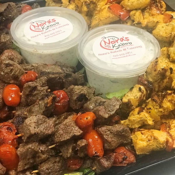 Noras Kabob catering tray with beef and chicken kabobs, roasted tomatoes, and two containers of sauce.
