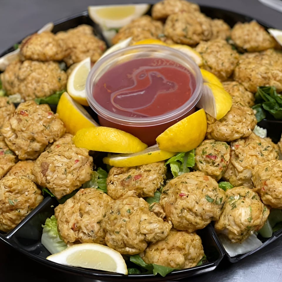 A platter of golden-brown crab cakes with lemon wedges, lettuce, and a cup of red sauce.