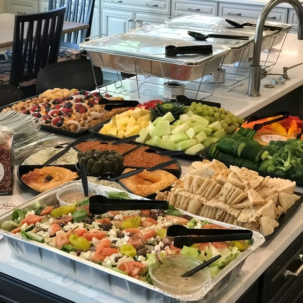 A buffet spread of Mediterranean food, including salad, hummus, pita bread, fruit, and pastries.