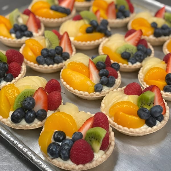 Rows of colorful fruit tarts with blueberries, kiwi, strawberries, raspberries, and peaches on a tray.