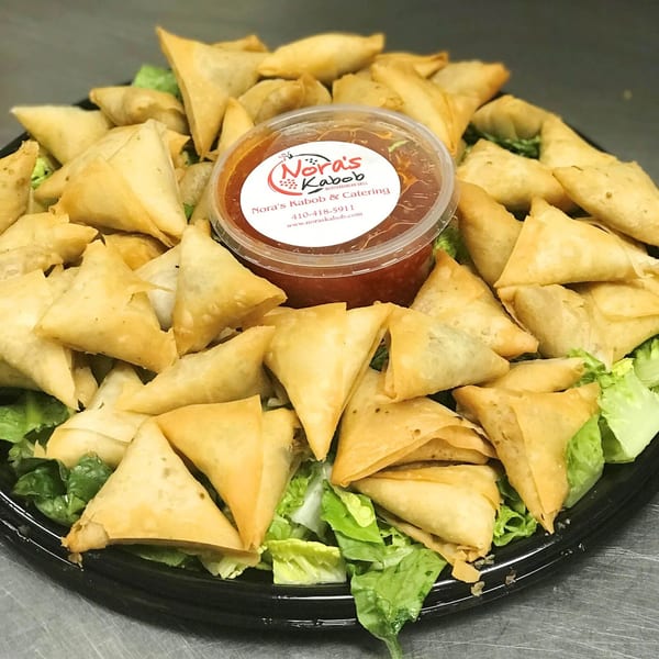 A platter of golden-brown samosas on a bed of lettuce, with a container of Nora's Kabob sauce.