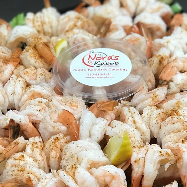 Close-up of Nora's Kabob shrimp platter with seasoning, lemon wedges, and a branded sauce container.